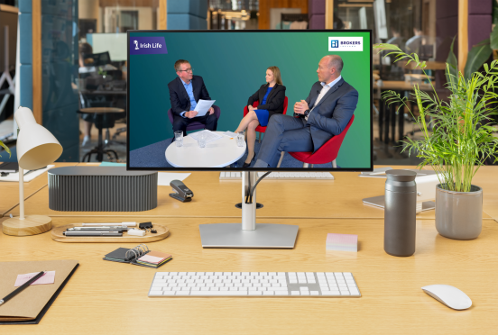A screen shows two men and one woman in business dress sitting around a small table while talking