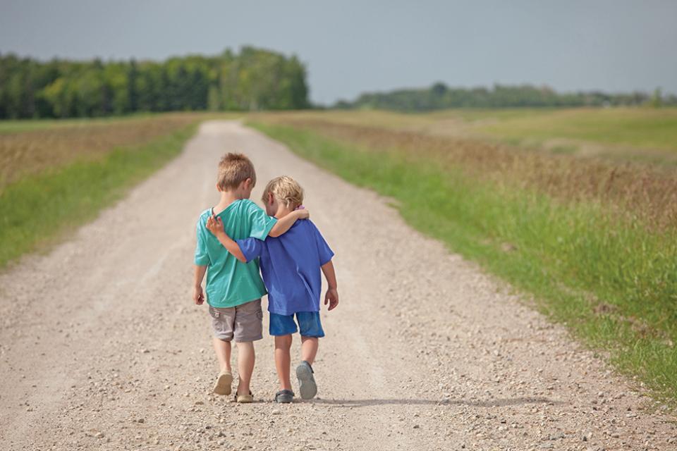 Two small children walk barefoot down a lane with their arms over one another's shoulders