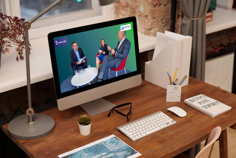 A screen on a desk showing two men and a woman sitting around a table in conversation