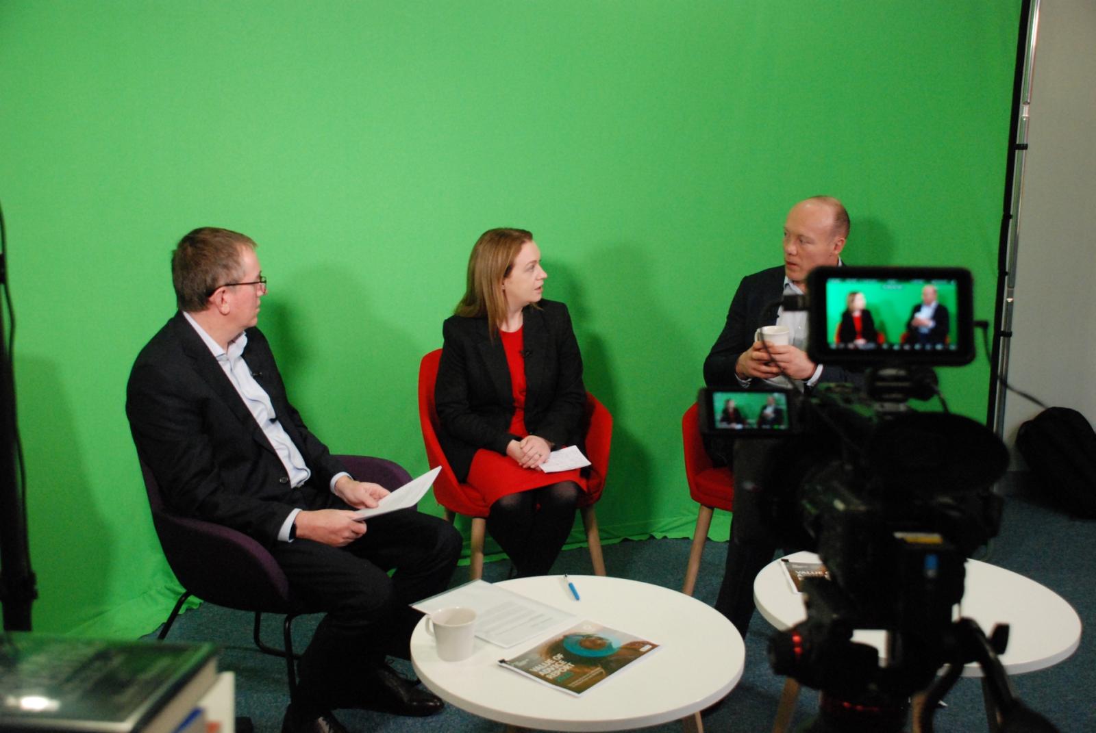 Two men and a woman sitting around a table in front of a green screen